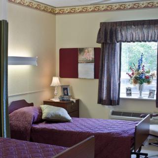 A modest nursing home room featuring two beds, a lamp, and a window with curtains.