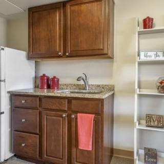 A small kitchen area featuring wooden cabinets, a granite countertop with a sink, open shelves, and a white refrigerator.