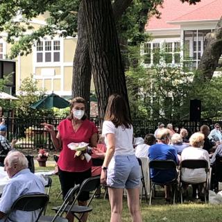 A community gathering outdoors with people seated at tables in a park-like setting.