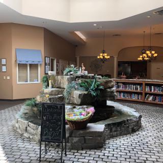 A serene indoor lobby featuring a decorative fountain surrounded by plants and bookshelves.