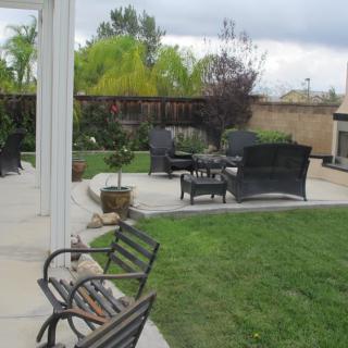 A backyard with a patio featuring outdoor seating and a fireplace, surrounded by a green lawn and garden.