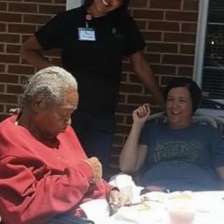 An elderly person sitting outside with a caregiver and another individual, engaged in conversation on a sunny day.