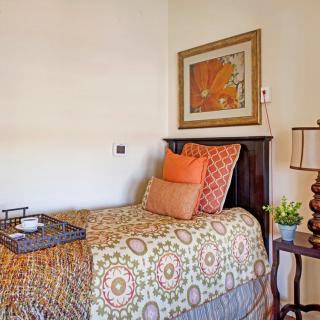 A cozy bedroom with a single bed adorned with colorful pillows and blanket, featuring a framed artwork on the wall and a lamp on the side table.