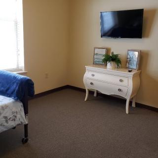 A minimalist bedroom with a mounted television, a white dresser adorned with frames and a plant, and natural light from the window.