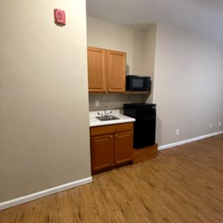 A small kitchen area in an empty apartment with wooden cabinets, a microwave, and a sink, featuring hardwood flooring.