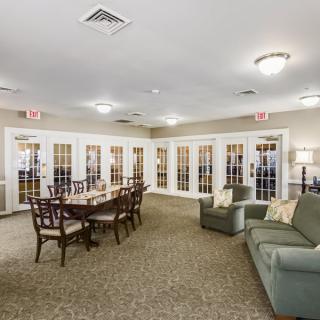A cozy meeting room with a large dining table, chairs, and a sofa, featuring bright lighting and carpeted flooring.