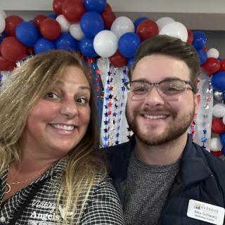 Two individuals posing for a cheerful photo at a festive event with patriotic-themed decorations.