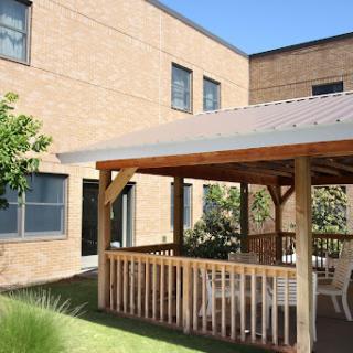 A cozy gazebo surrounded by greenery, adjacent to a modern building under a clear blue sky.