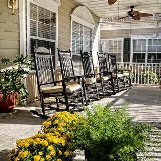 A cozy front porch with several rocking chairs lined up, surrounded by potted plants and flowers.