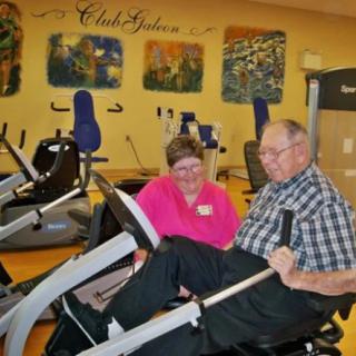 An elderly man using workout equipment with assistance from a healthcare professional in a gym setting.