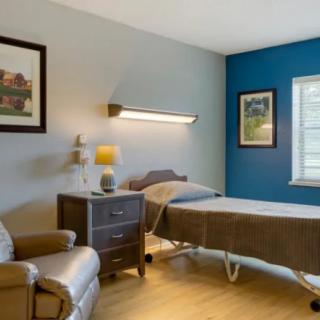 A cozy hospital room featuring a bed, armchair, and bedside table with a lamp, adorned with paintings on the walls.