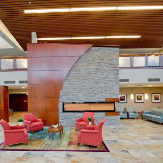 A modern lobby with red chairs surrounding a fireplace, featuring a stone wall and a wooden ceiling.