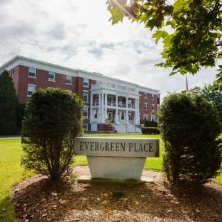A grand brick building with white columns stands behind a sign reading 'Evergreen Place', surrounded by trees and greenery.