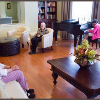 Elderly individuals sitting in a living room with a piano and bookshelves.
