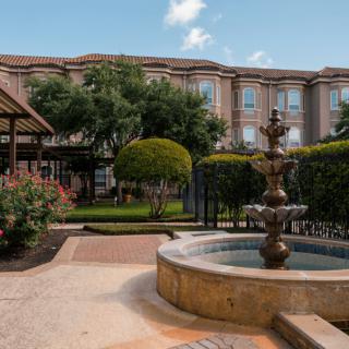 A scenic courtyard featuring a fountain surrounded by well-maintained gardens and a large building in the background.