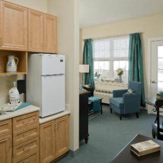 A cozy apartment interior featuring a small kitchen area and a bright living space with blue chairs and drapes.