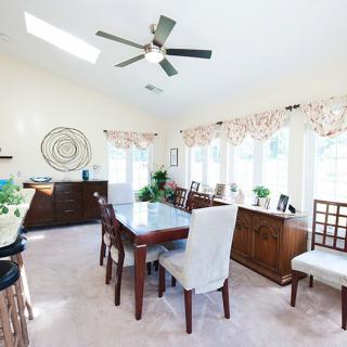 A bright dining room with a large table, surrounded by chairs and decorated with indoor plants, featuring a ceiling fan and ample natural light from the windows.