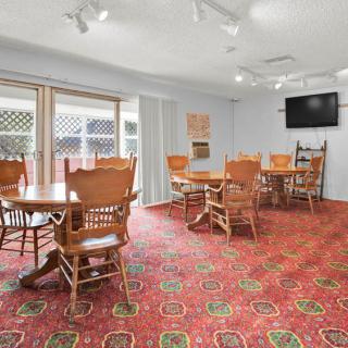 A dining room with wooden tables and chairs, featuring a patterned carpet and a wall-mounted TV.