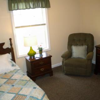 A cozy bedroom with a bed, nightstand, and an upholstered chair by the window.