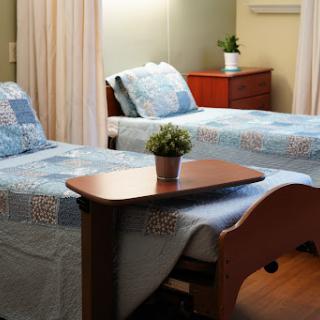 A cozy hospital patient room featuring two beds with blue and white quilts, a small table with a potted plant, and warm wooden flooring.