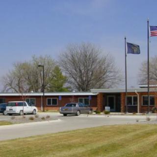 A small brick building with flags and parked cars in front, surrounded by trees and a well-kept lawn.