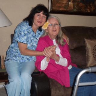 An elderly woman and a caregiver sitting together on a couch in a home setting.