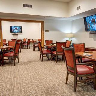 An empty restaurant dining area with wooden tables and chairs, carpeted flooring, and TV screens mounted on the walls.