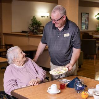 A caregiver warmly serves a meal to an elderly resident in a comfortable dining setting.