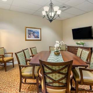 A well-furnished conference room featuring a long wooden table surrounded by chairs, a chandelier above, and a TV mounted on the wall.