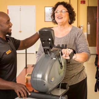A woman uses an exercise bike while a trainer assists her in the gym.