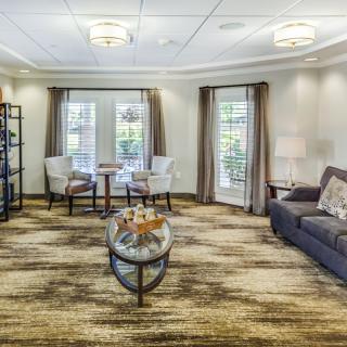 A cozy living room with modern decor, featuring a sofa, chairs, and a coffee table centered on a patterned rug. Large windows with curtains allow natural light to fill the space.