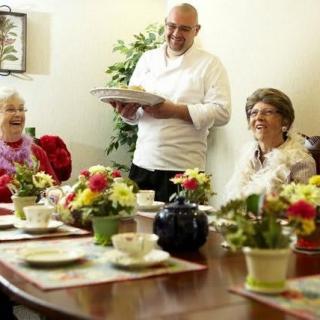 A chef serving a meal to elderly individuals gathered around a table adorned with flowers.
