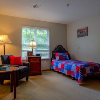 A cozy bedroom featuring a twin bed with a colorful quilt, a comfortable chair, and a warm lamp by the window.