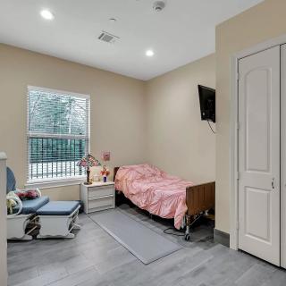 A cozy bedroom with a single bed featuring pink bedding, an armchair by the window, a nightstand with a lamp, and a wall-mounted TV.