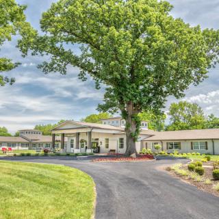 A scenic view of a building with a curved driveway and large tree in the foreground under a partly cloudy sky.