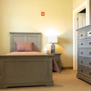 A cozy bedroom featuring a single bed, matching dresser, and lamp.