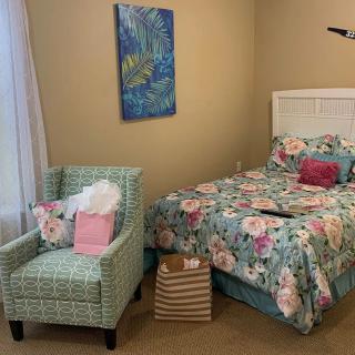 A cozy bedroom featuring a bed with floral bedding and a matching armchair, decorated with a colorful painting on the wall.