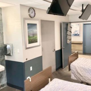 A clean hospital room with two beds, a clock, and artwork on the walls.