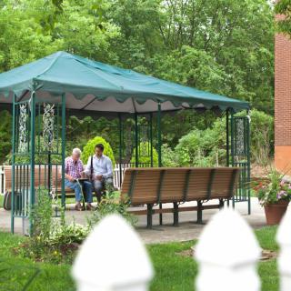An elderly couple sitting and relaxing in a gazebo surrounded by lush greenery.