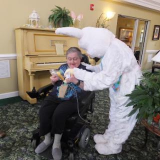 A cheerful Easter celebration at an assisted living facility, where a person in a bunny costume engages with a resident in a wheelchair.