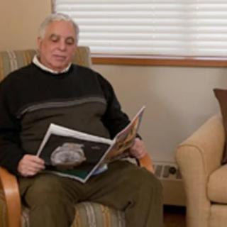 An elderly man sitting in an armchair, reading a magazine indoors.