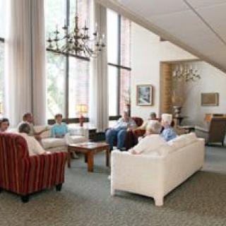 A group of people conversing in a spacious living room with large windows and comfortable seating.