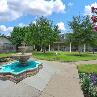 A beautiful garden featuring a central fountain surrounded by trees and flowers under a bright blue sky.
