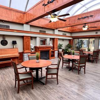 A spacious, well-lit interior with wooden furniture, featuring tables, chairs, a piano, a fireplace, and decorative plants.