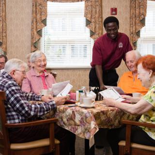 A group of senior citizens enjoying a meal together in a care home, assisted by a staff member.