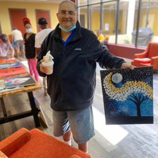 A person proudly displays a painting they created while holding a cup inside an art studio.