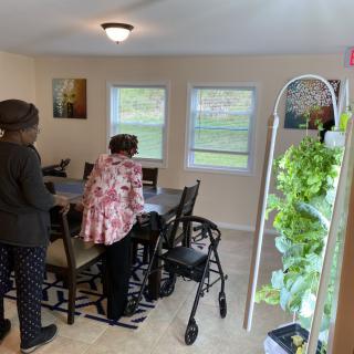 Two individuals standing in a dining area with a walker and indoor plants visible.