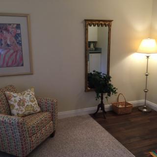 A cozy living room corner featuring a patterned chair, floor lamp, decorative mirror, artwork on the wall, and a small plant.