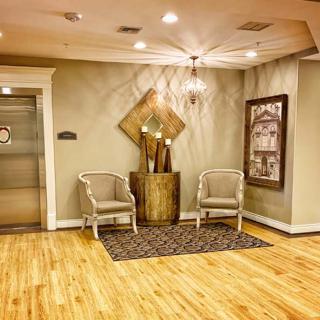 A warmly lit lobby area featuring two chairs, a central decorative vase, and an elevator.