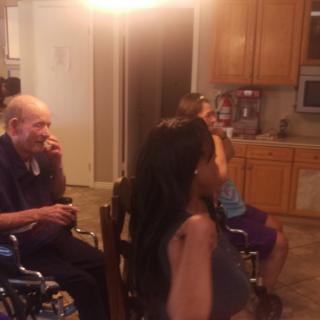 A family gathering in a living room with an elderly man in a wheelchair and others seated nearby.